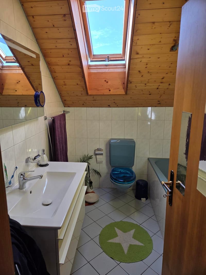 Bathroom with a white sink, silver faucet, blue toilet, bathtub, and a green bath mat with a white star design. Wooden ceiling and tiled walls.