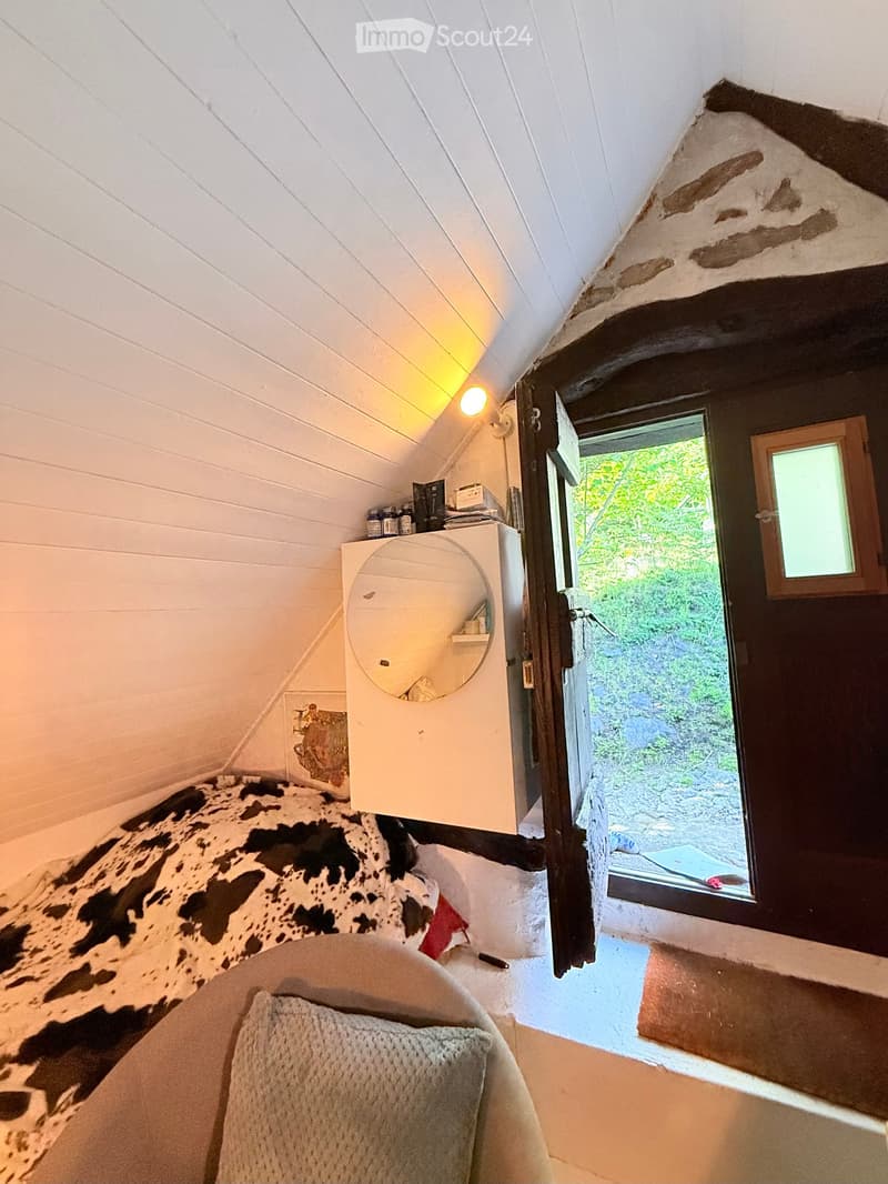 Cozy attic bedroom with slanted wooden ceiling, cowhide bedspread, round mirror, and open wooden door leading to a green outdoor view.