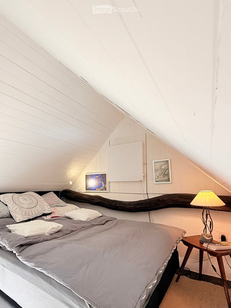 Cozy attic bedroom with slanted wooden ceilings, a large bed with grey bedding and patterned pillows, a wooden nightstand with a lamp, and two picture frames on the wall.