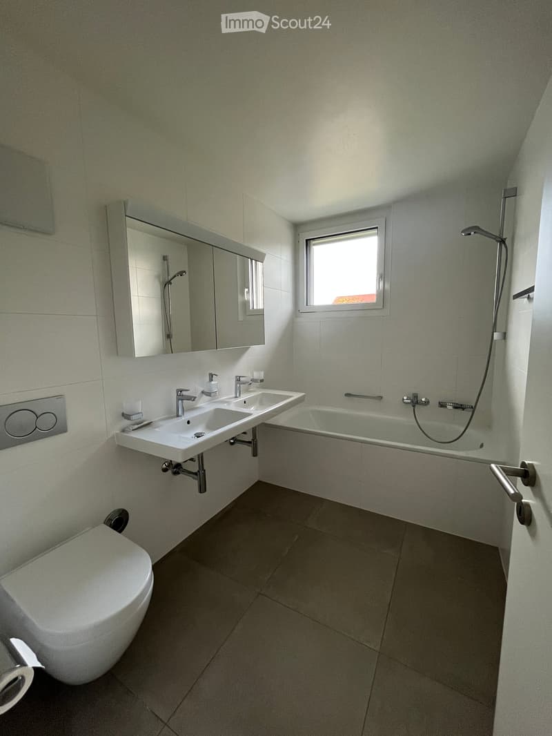 Modern bathroom with a toilet, double sink, bathtub, large mirror, and window providing natural light.