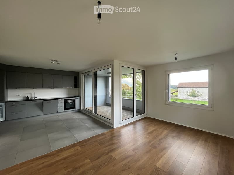 Open-plan living and kitchen area with wooden flooring, grey kitchen cabinets, and large windows offering a view of the countryside.