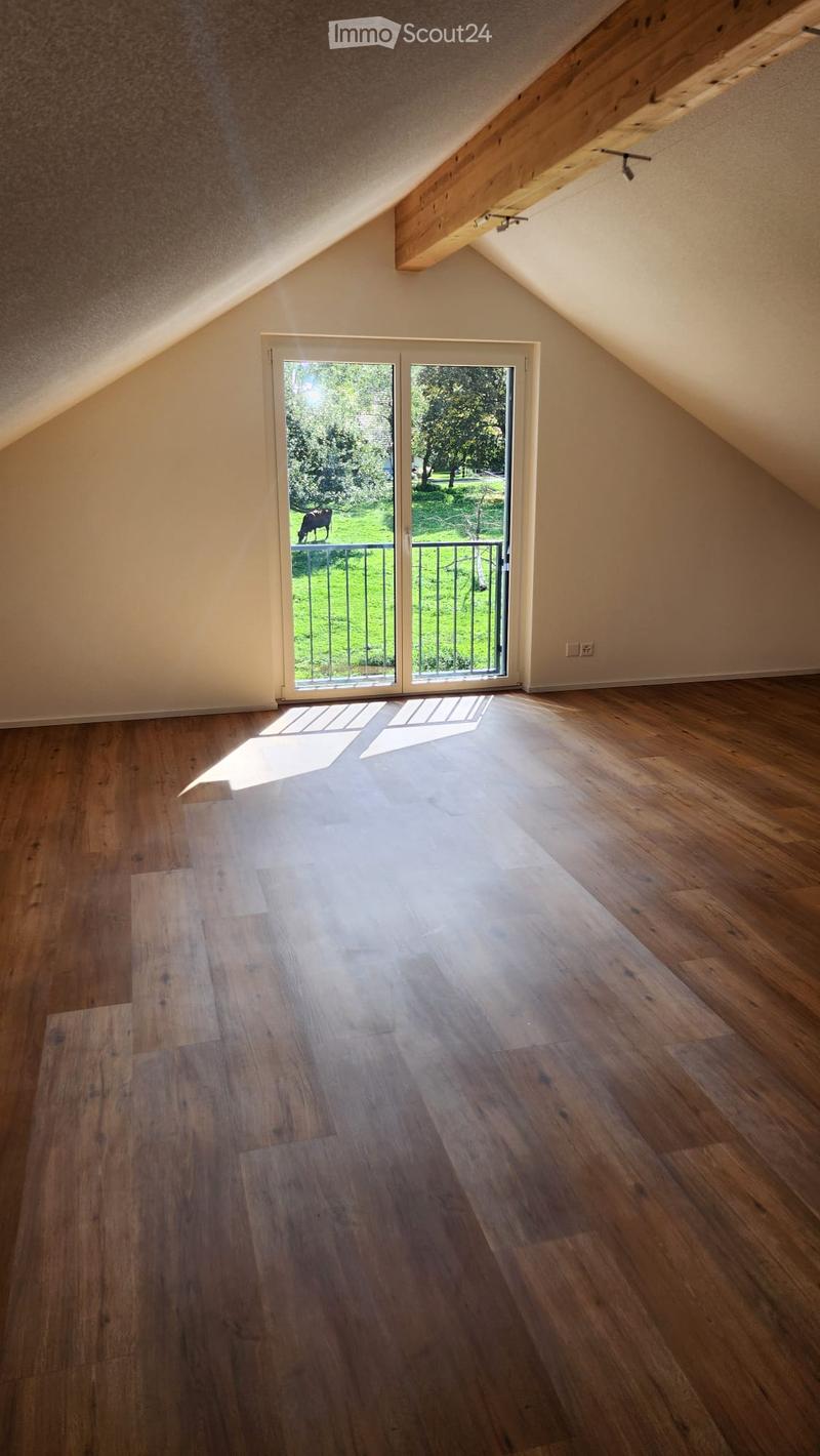 Empty attic room with wooden flooring, slanted ceilings, and a large sliding glass door leading to a balcony with a view of a green lawn and trees.