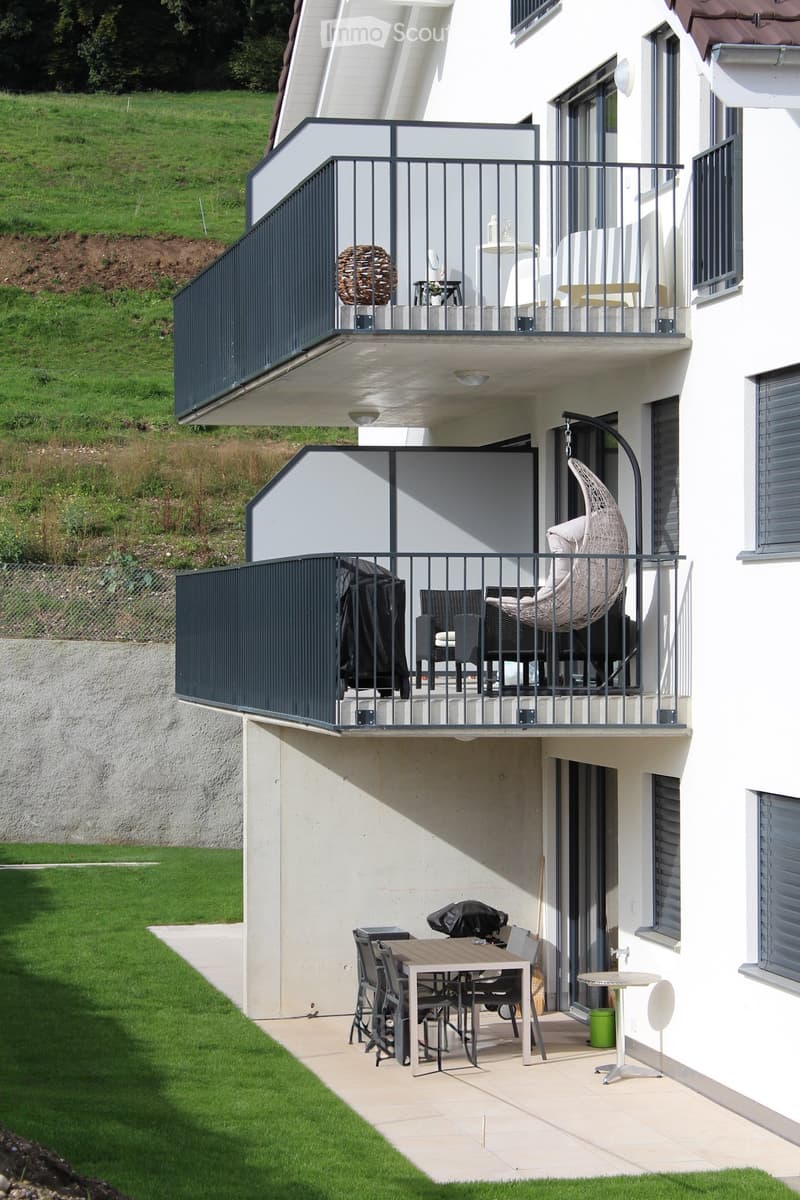 Balcony with a table, chairs, and a wicker hanging chair, overlooking a grassy area.