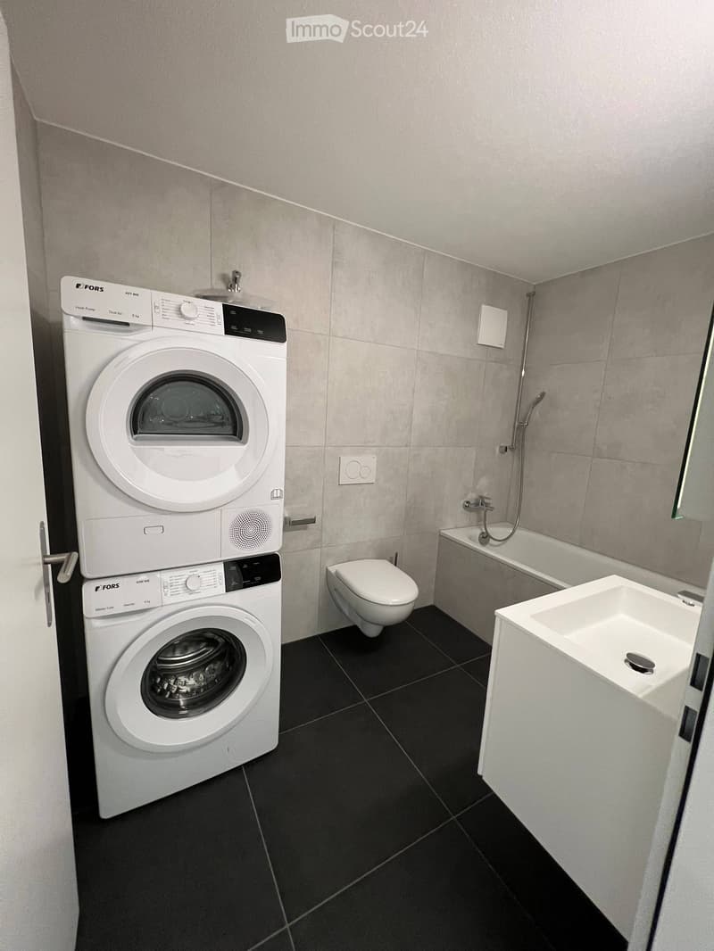 Modern bathroom with black tiled floor, white washing machine and dryer stacked, toilet, bathtub, and sink.