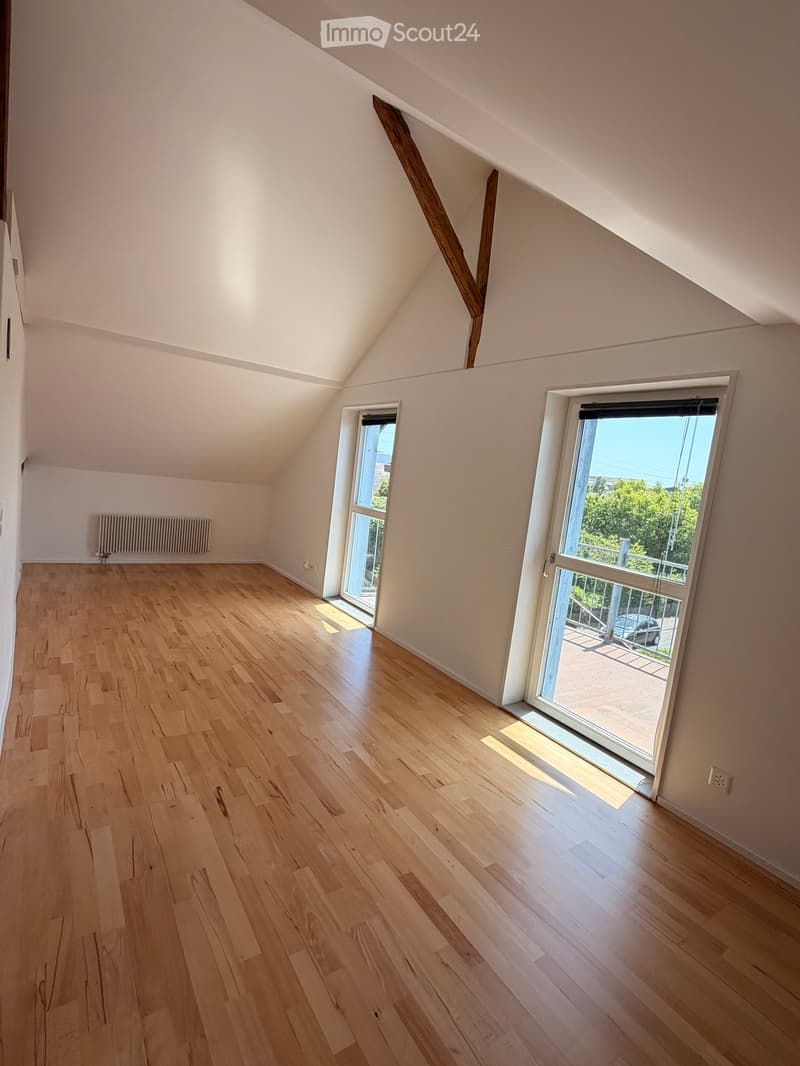 Empty attic room with wooden flooring, white walls, and large windows offering natural light. The room features exposed wooden beams and a radiator on one wall.