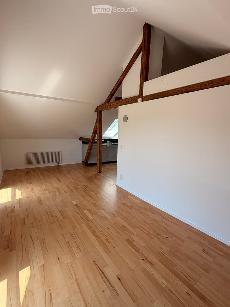 Empty attic room with wooden floors, white walls, exposed wooden beams, and a small kitchen area visible in the background.