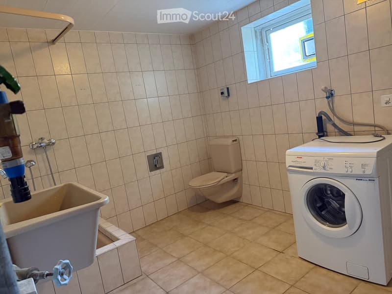 Bathroom with beige tiled walls and floor, featuring a toilet, a washing machine, and a bathtub.