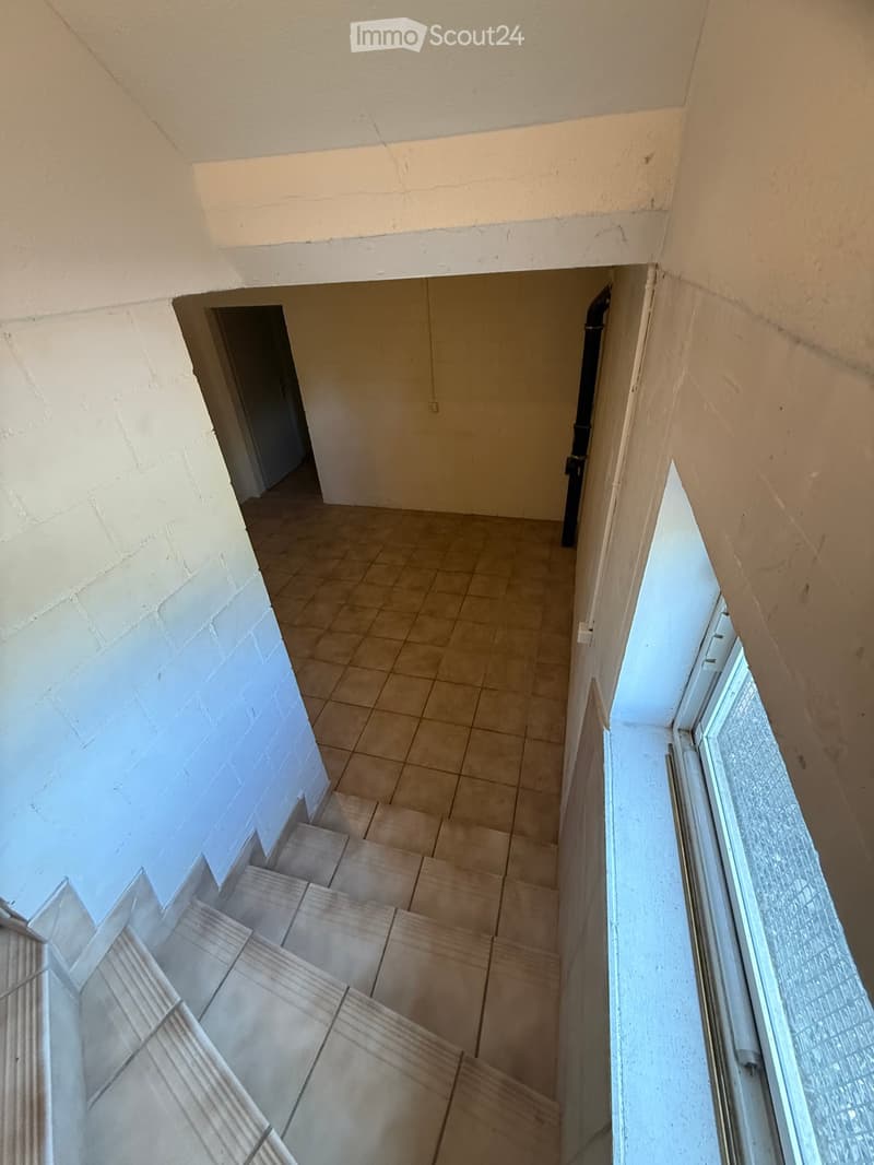A narrow hallway with tiled flooring, white walls, and a staircase leading upwards. There is a door on the left and a window on the right.