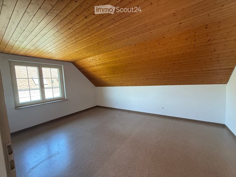 Empty attic room with wooden slatted ceiling, white walls, carpeted floor, and a single window with white frame and multiple panes.