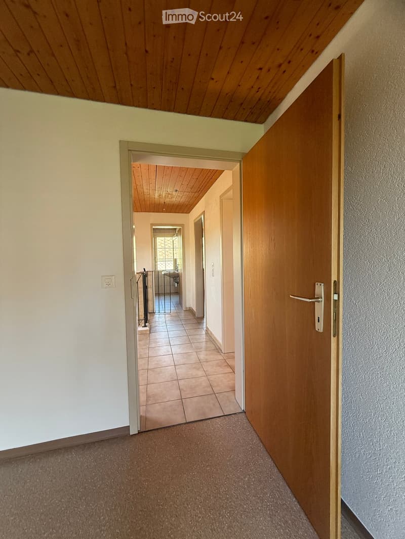 A narrow hallway with wooden doors, tiled floor, and a wooden ceiling. The hallway leads to another room with a visible staircase and railing.
