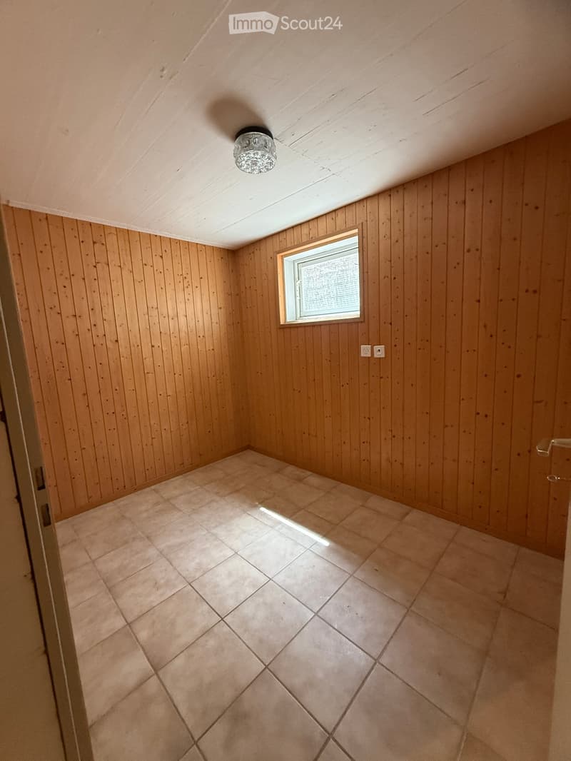 Empty room with wooden walls, tiled floor, and a single window letting in natural light. A ceiling light fixture is visible.