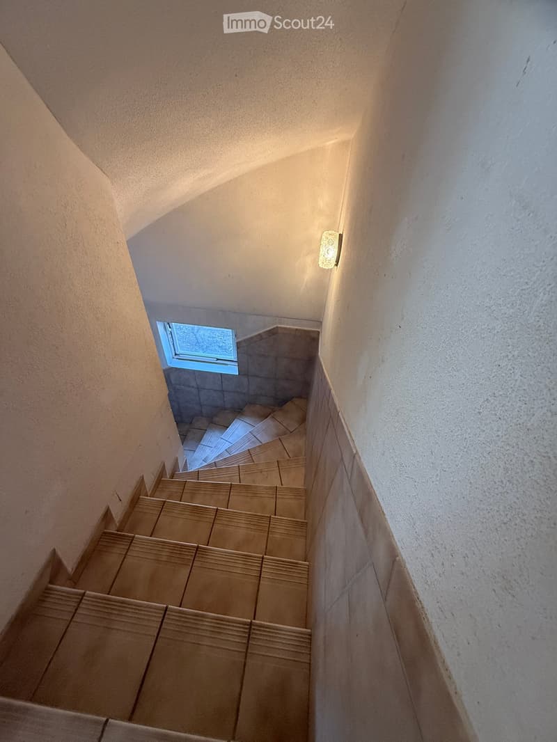 Staircase with beige tiled steps and walls, small window at the top, and a wall-mounted light fixture on the right side.