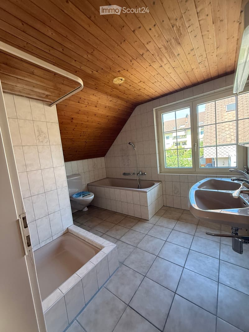 Bathroom with a wooden slanted ceiling, tiled walls and floor, bathtub, toilet, and double sink.