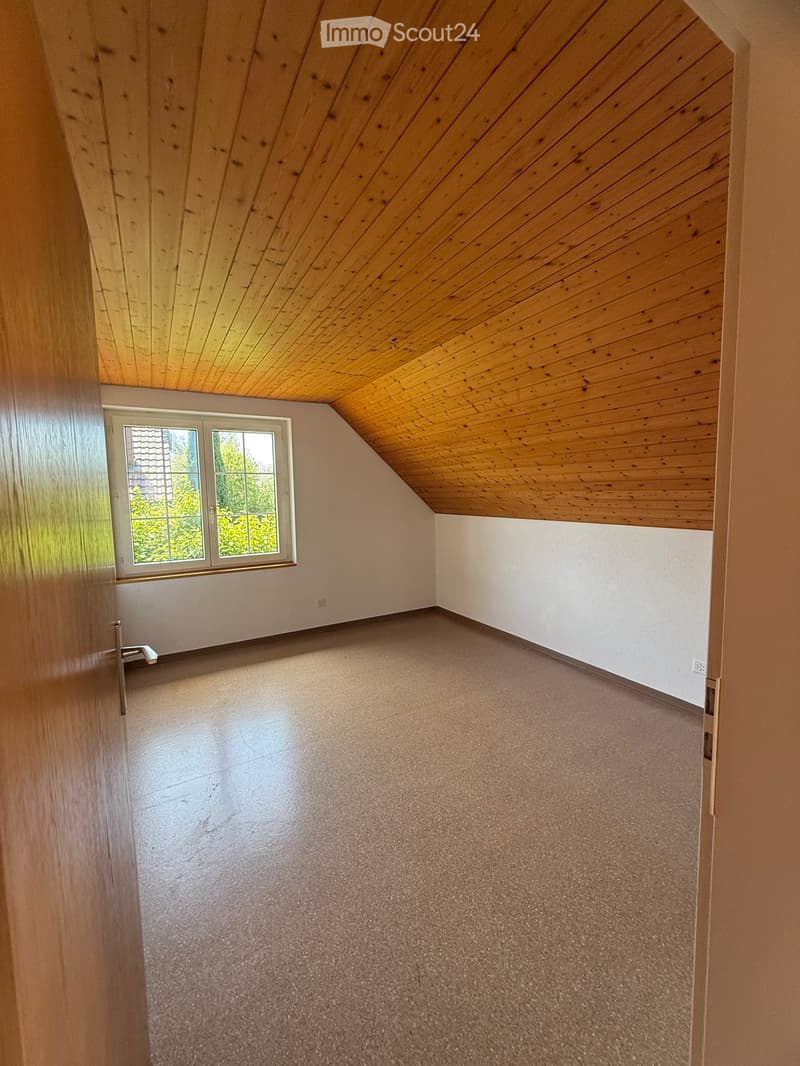 Empty room with wooden slanted ceiling, carpeted floor, and a window with white frame and glass panes.