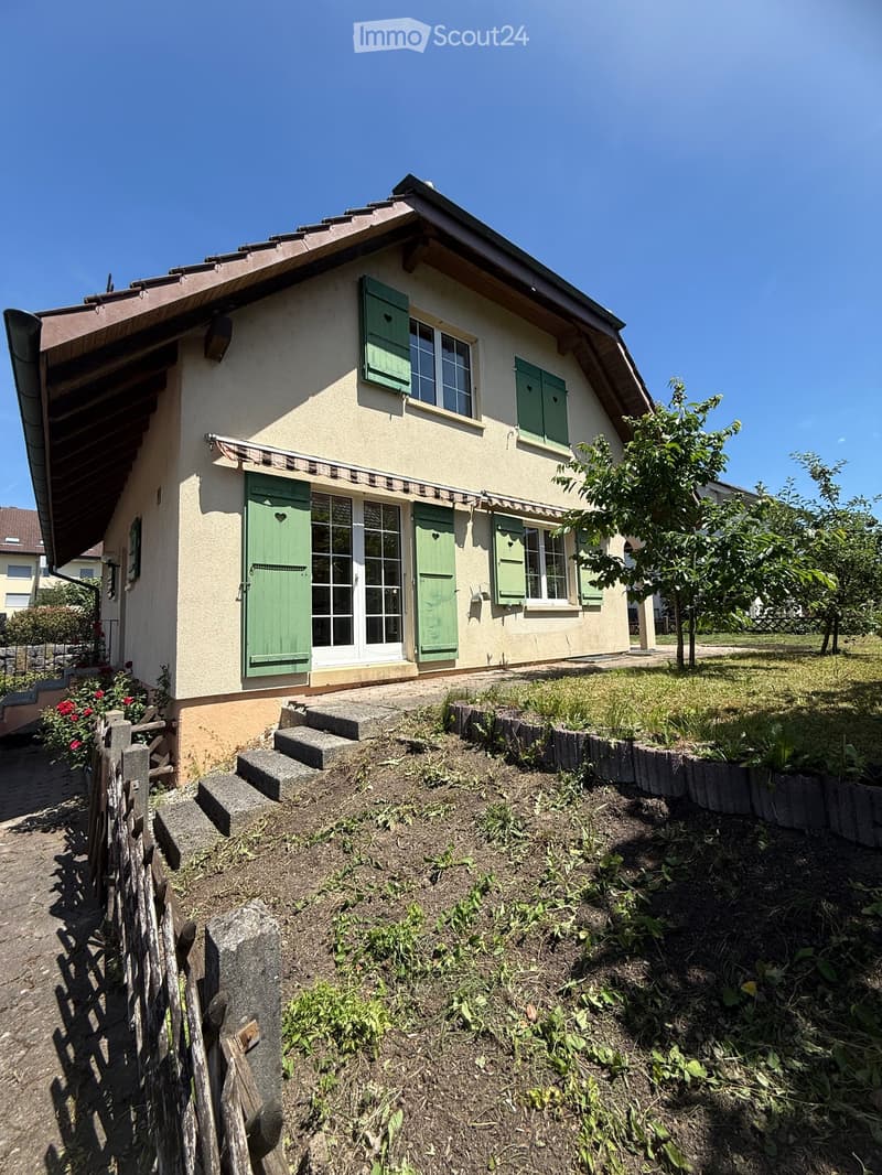 Two-story house with beige walls, green shutters, and a sloped roof. The entrance has steps leading up to a door with glass panels, flanked by green shutters. There is a small garden area with a wooden fence and some plants.