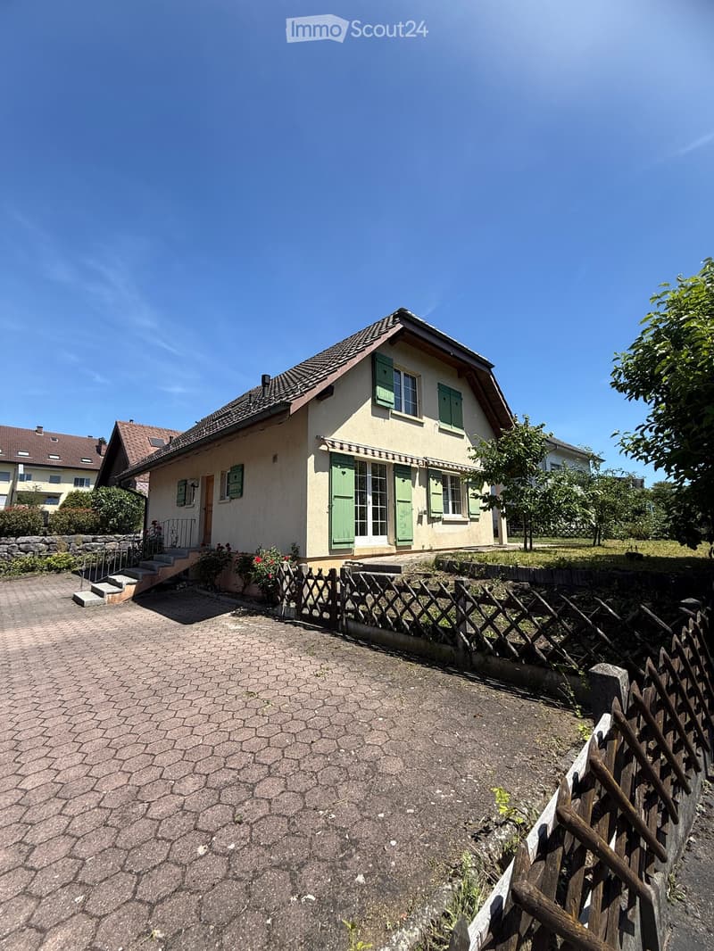 Two-story house with beige walls, green shutters, and a tiled roof. The house has a small front porch with steps leading up to the entrance, surrounded by a wooden fence and some greenery.