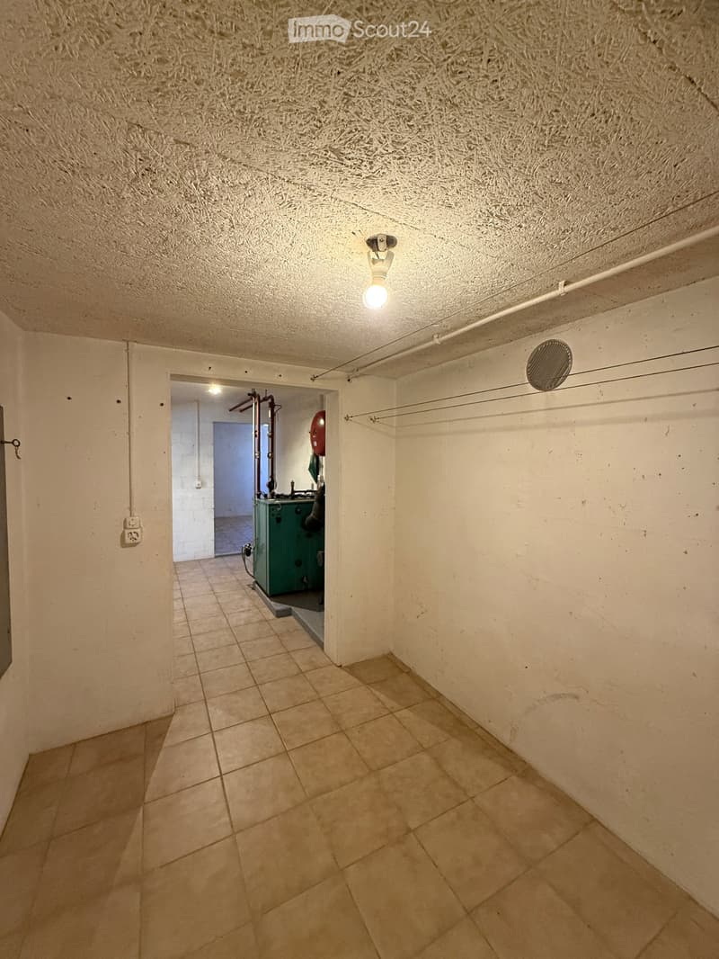 Empty hallway with tiled floor, white walls, and a textured ceiling. A small room with a green machine is visible through an open doorway.