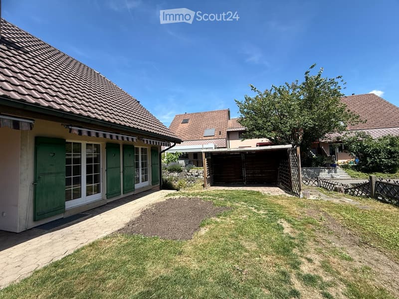 A well-maintained garden area with a paved pathway leading to a house with green shutters and a striped awning. The house has a tiled roof and is surrounded by other residential buildings. There is a wooden garage and a small stone staircase leading up to