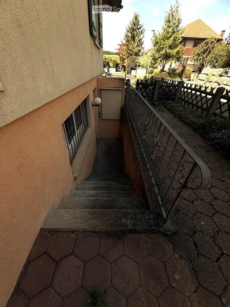 A narrow concrete staircase leading up from a paved area, with a metal railing on one side and a wooden fence on the other. The staircase is bordered by a beige building wall on the left and a small garden area on the right.