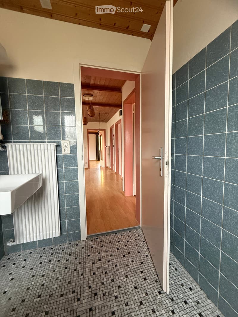 An open doorway leading to a hallway, bathroom wall with blue tiles, white tile floor, white sink with white radiator below it, electrical outlets on the wall, and hallway has wooden floors and pink walls.