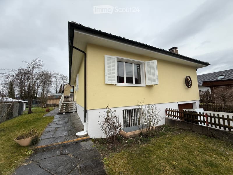 Raised ground floor, yellow and white painted house, white shutters, gravel pathway, fenced garden, chimney, metal railings on staircase