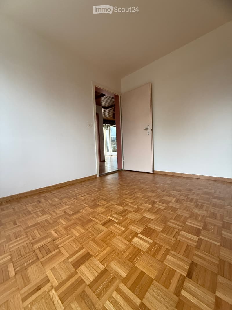An empty room with wooden floors, white walls, a doorway, and a window in the back wall.