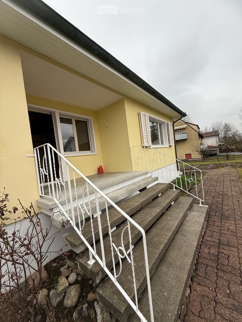Yellow house with white railings, concrete steps leading up to the entrance, red object near the door