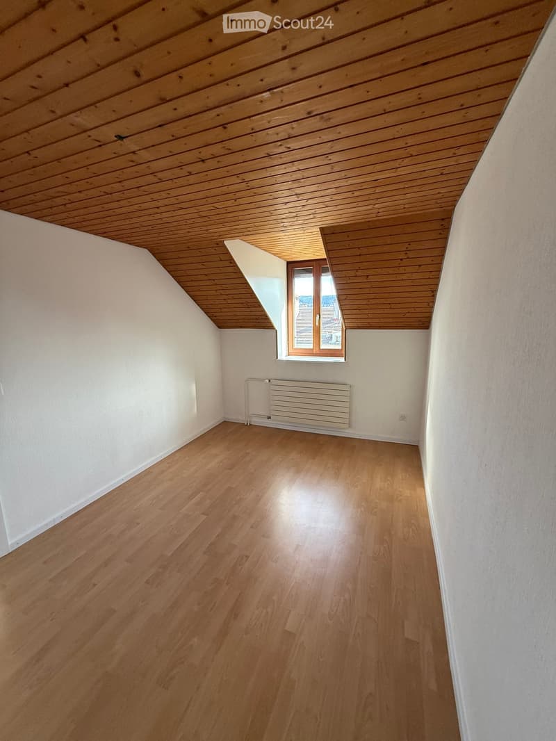 Empty room with wooden flooring, white walls, and a slanted wooden ceiling. A window with a view and a radiator are visible.