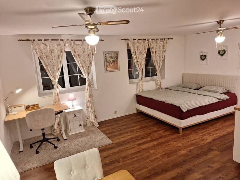 Cozy bedroom with wooden flooring, a large bed with striped bedding, a desk with a chair, a nightstand with a lamp, and two ceiling fans with lights.