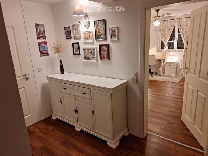 A hallway with a wooden floor, a white cabinet with a bottle on top, several picture frames on the wall, and an open doorway leading to a room with a chair, table, and lamp.