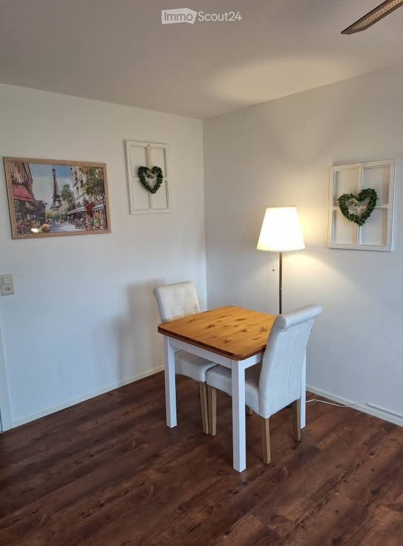Dining area with wooden floor, white walls, a small wooden dining table with two white chairs, a floor lamp, and decorative wall art including a framed picture and a heart-shaped wreath.