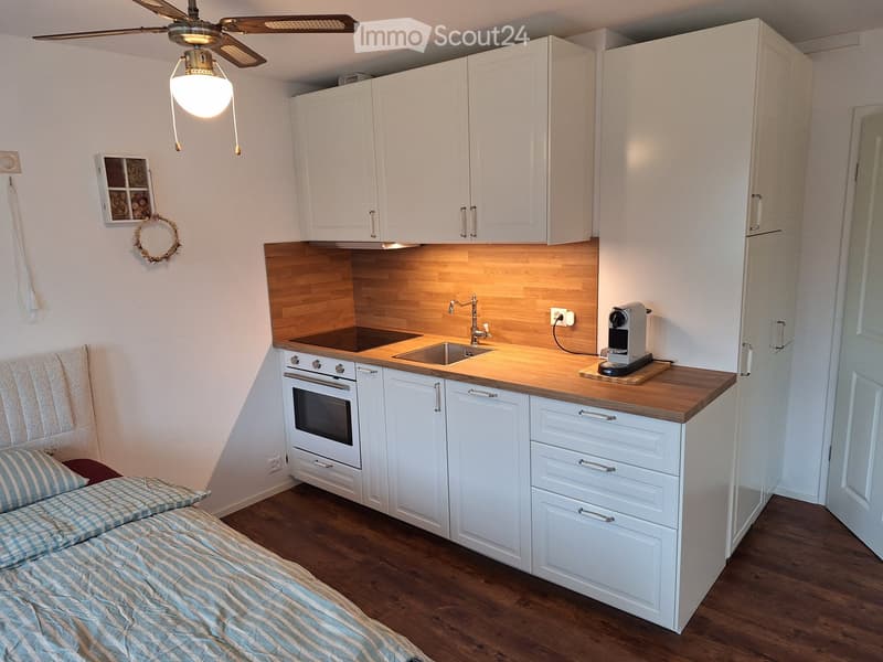A compact studio apartment with a bed on one side and a kitchen area on the other, featuring white cabinets, a wooden countertop, and a ceiling fan with light.