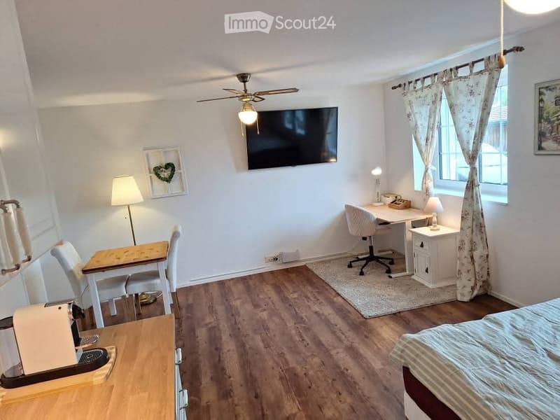 Cozy room with a bed, desk, and seating area. Features a television mounted on the wall, a ceiling fan with light, and a window with floral curtains.