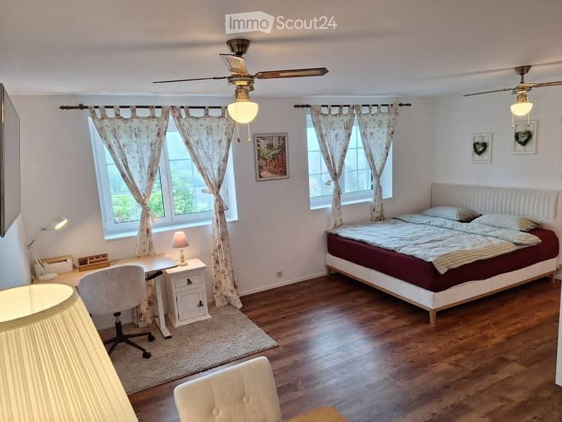 Cozy bedroom with wooden flooring, large bed with red and white bedding, desk with chair, and two ceiling fans with lights.