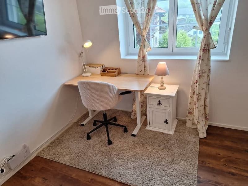 A cozy, well-lit room with a wooden desk, ergonomic chair, table lamp, nightstand, and floral curtains. The room features a combination of wooden flooring and a soft, light-colored rug.