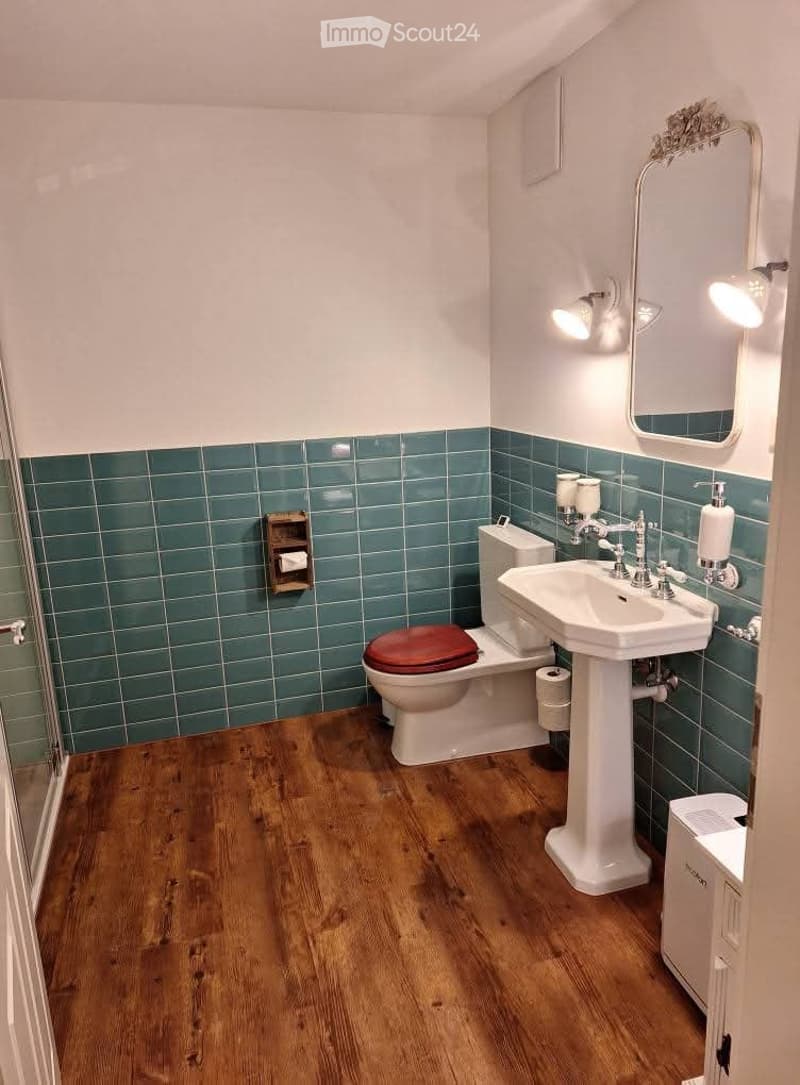 Bathroom with teal tiled lower walls, wooden flooring, a toilet with a red seat, a pedestal sink, a mirror with a decorative frame, and a wooden shelf holding toilet paper.