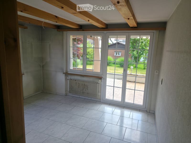 Empty room with exposed wooden beams, tiled floor, large sliding glass doors leading to a patio, radiator, and a small shelf.