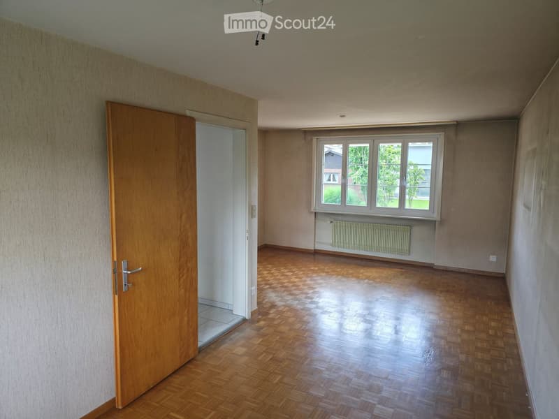 Empty room with wooden flooring, large window, radiator, and open wooden door leading to another space.