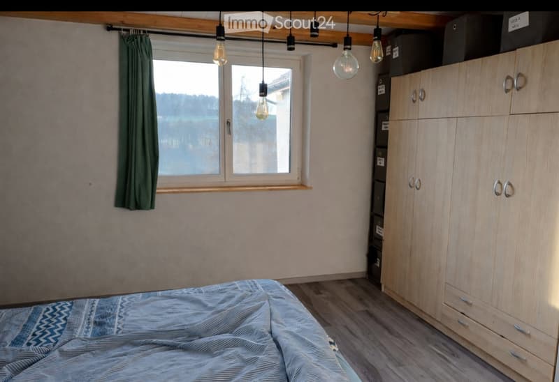 Cozy bedroom with a wooden wardrobe, a neatly made bed, and a window with green curtains. Exposed wooden beams on the ceiling and pendant lights add a rustic touch.