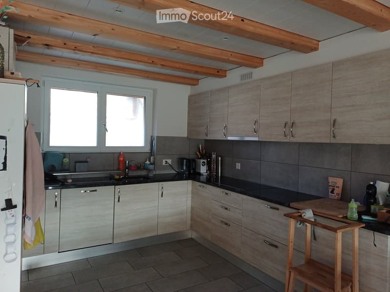 Modern kitchen with light wood cabinets, stainless steel appliances, and a black countertop. Features include a sink, coffee maker, cutting board, and a small wooden stool.
