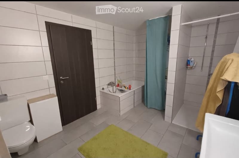 Modern bathroom with white tiled walls and floor, featuring a toilet, bathtub, shower area with a blue curtain, and a yellow bath mat on the floor.