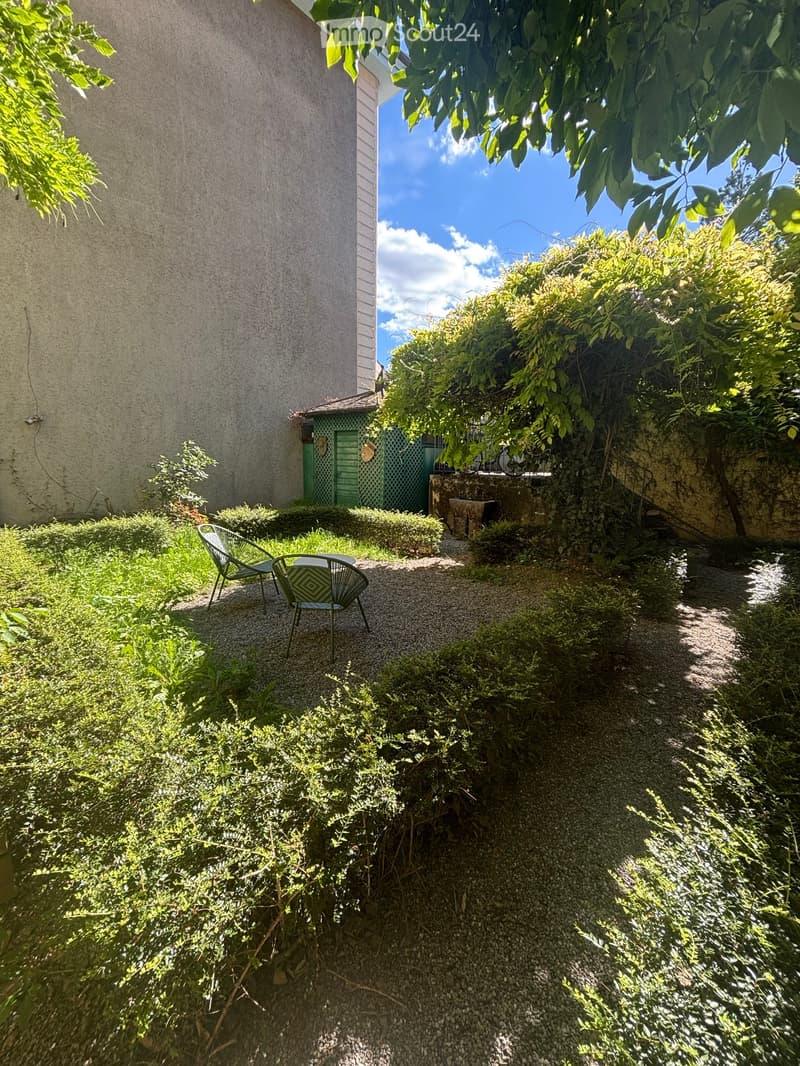 A small, lush garden area with gravel pathways, greenery, and a few outdoor chairs. A building with a green door is visible in the background.
