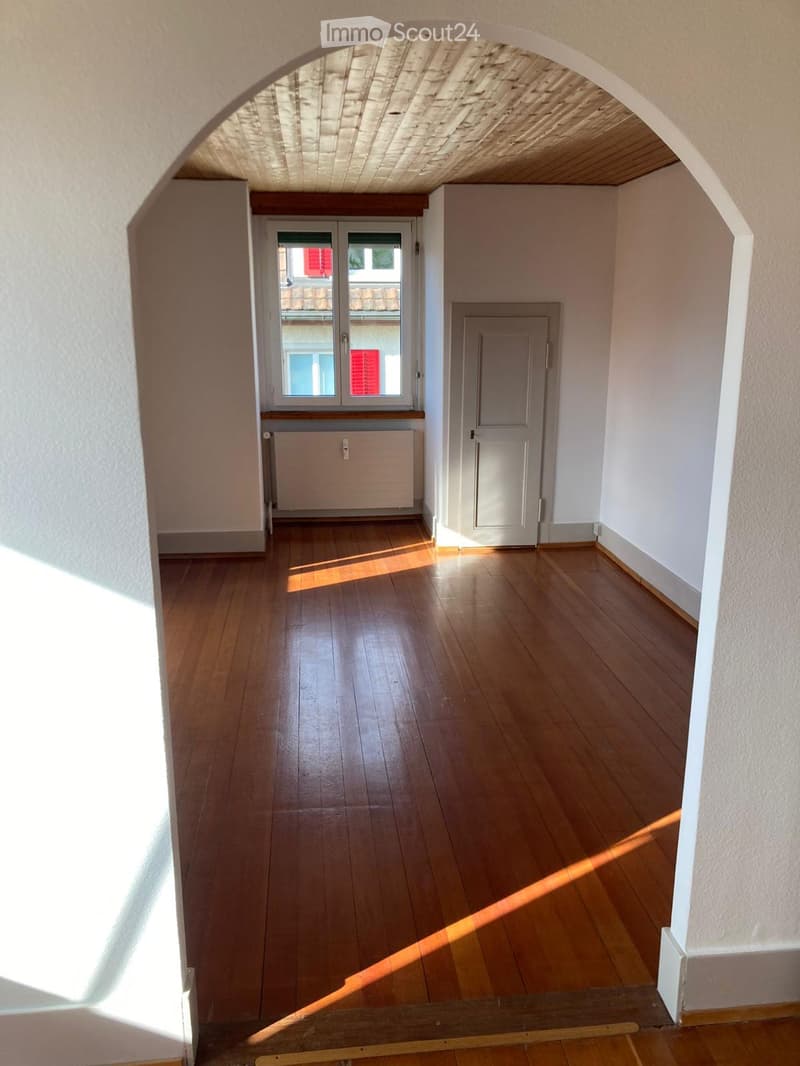 Empty hallway with wooden flooring, white walls, a window with a view of a building, and a closed door.