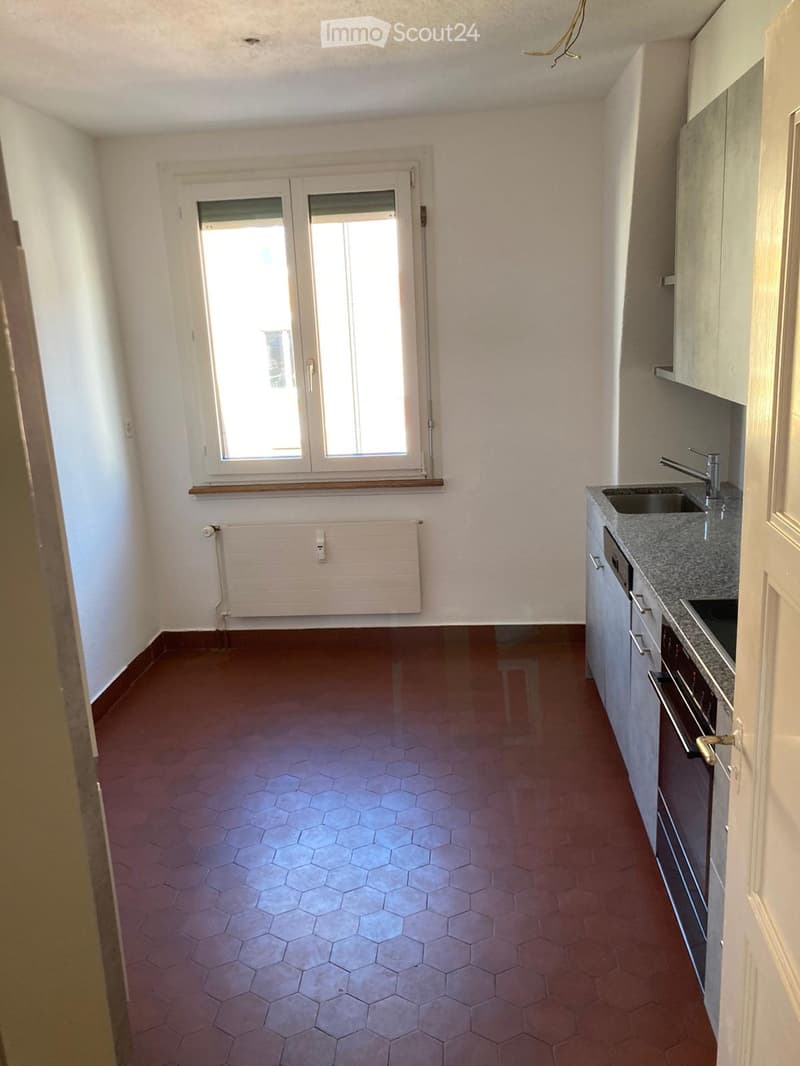 Empty kitchen with modern cabinets, a sink, and an oven. The floor is tiled in a hexagonal pattern, and there is a window with a radiator below it.