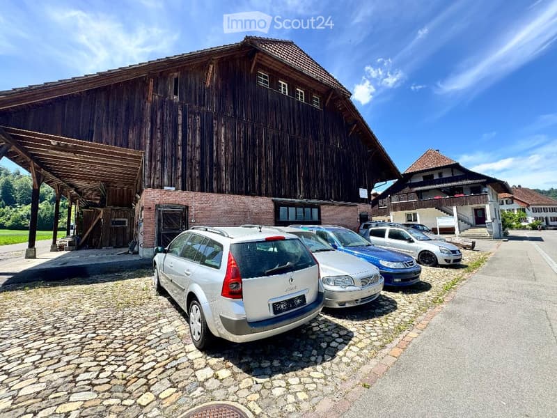 Large wooden barn with brick base, multiple parked cars in front, clear blue sky, surrounding houses visible.