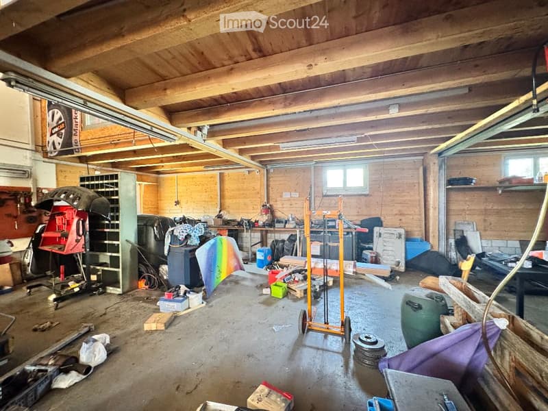 A spacious garage with exposed wooden beams, concrete floor, and various tools and equipment, including a workbench, shelves, and car parts.