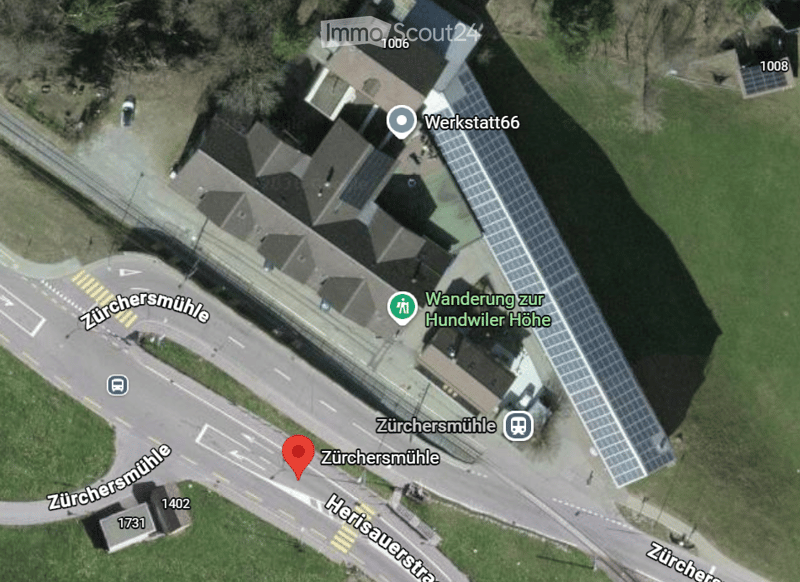 Aerial view of a building complex with multiple structures, solar panels on the roof, and surrounding green areas. Nearby roads and pathways are visible.