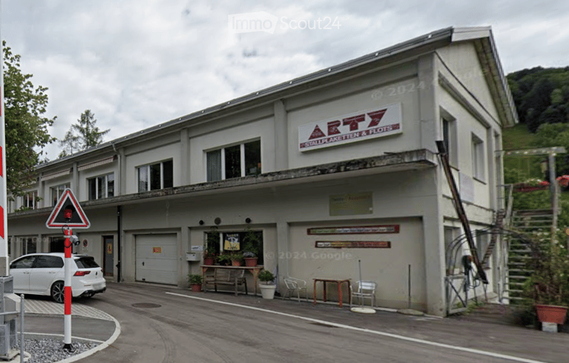 Two-story building with a garage, signage reading "ARTY Stallplaketten & Plots", and outdoor seating area with tables and chairs.