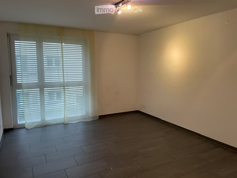 Empty room with wooden flooring, white walls, large window with blinds and curtains, and recessed ceiling lighting.