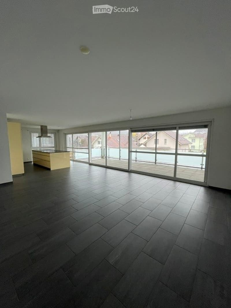 Empty room with dark wooden flooring, large sliding glass doors leading to a balcony, and a small kitchen area visible in the background.
